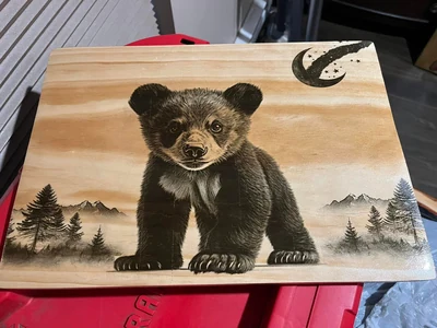 Bear cub with crescent moon, pines and mountains on a pine board